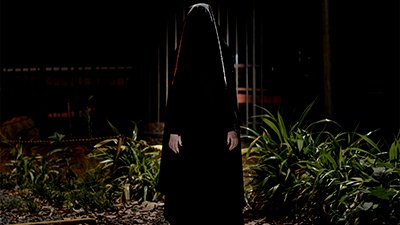 Fotografia de uma cena do filme A triste figura, de Calebe Lopes. BA, 2018, FICÇÃO, 18 min. Na noite escura, uma pessoa com véu preto sobre a cabeça e uma túnica larga embaixo, também preta, de manga comprida, tem as mãos brancas, ao lado das coxas. Uma luz fria, por trás contorna a cabeça e o véu até abaixo dos ombros e as plantas, nas laterais da estranha encapuzada. Ao fundo, um portão e grades de ferro estão na penumbra.
