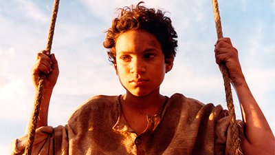 Fotografia de uma cena do filme Abril despedaçado, direção de Walter Salles. RJ, 2001, FICÇÃO, 100 min., Cor, H264 Do peito para cima, o então menino, Ravi Ramos Lacerda, de pele clara, cabelos castanhos cacheados em pequeno Black, olhos em igual tonalidade, nariz arredondado, lábios finos fechados em um bico. Está entre duas cordas, as quais segura uma em cada uma das suas mãos manchadas em tom escuro, na direção das orelhas. Usa camisa amarronzada, larga de botões, gola careca e mangas compridas arregaçadas na altura dos cotovelos flexionados. Tem a metade da sua face direita iluminada pelo sol, seu meio olhar fixo, atravessado na mesma direção, ao fundo, desfocado, céu azul claro com algumas rasas nuvens.