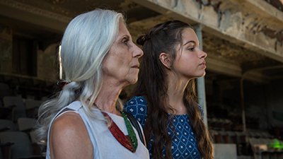 Fotografia de uma cena do filme Guerra de algodão, de Marília Hughes e Cláudio Marques - BA, 2019, FICÇÃO, 82 min. Duas mulheres brancas, de perfil direito, do busto para cima. A primeira, à esquerda, Maria, olha fixo à frente, tem cabelos lisos grisalhos ao longo das costas, presos para trás, nariz afilado e pele enrugada. Usa roupa clara, colar vermelho e verde. Ao lado, Dora, olhar decidido, nariz e queixo empinado, cabelos lisos longos, castanhos claro, com trança presa para trás e o restante solto, e pele lisa cintilante. Usa roupa de mangas, azul escura com detalhes triangular branco. O fundo desfocado, é um ambiente pouco iluminado com arquitetura antiga descascada, em tons pasteis, com poltronas enfileiradas, janela escura e teto rebaixado.