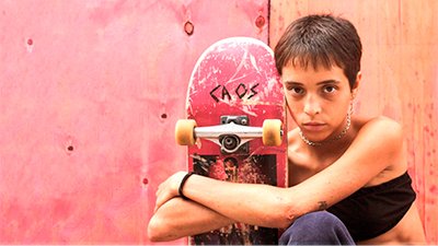 Fotografia de uma cena do filme Meu nome é Bagdá, de Caru Alves, RJ, 2019, Ficção, 100 min. Bagdá, uma jovem de 17 anos, magra, cabelos curtos, penteados para frente, usa gargantilha prata, top preto e calça azul. Está sentada na calçada abraçada ao skate vermelho em posição vertical. O rosto dela encosta no skate que tem escrito caos. Ela olha fixo para frente. Atrás dela tábuas de madeirite.