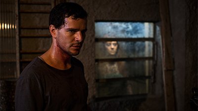 Fotografia horizontal de uma cena do filme Morto não fala, de Dennison Ramalho. RS, 2019, FICÇÃO, 110 min. À esquerda, quase de perfil direito, um homem, jovem, branco, de cabelos curtíssimos pretos, de olhar contraído, rosto suado, com uma mancha de sangue no pescoço. Ele usa uma camiseta cinza escuro. Atrás dele, um ambiente de pouca iluminação, com um espaldar de madeira, sobre uma parede branca crespa e à direita, uma janela de vidro suja. Olhando para dentro, sob uma luz no rosto, uma mulher séria, de cabelos ondulados, castanhos, na altura dos ombros, casaquinho marrom, sobre blusa branca.