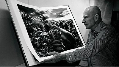 Fotografia de uma cena do filme O sal da terra, de Wim Wenders, Juliano Salgado. BR/FRA/ITA, 2014, DOCUMENTÁRIO, 110 min. Em preto e branco, Sebastião Salgado, folheia um dos seus livros. De perfil esquerdo, ele usa a cabeça raspada, tem as sobrancelhas brancas e está com uma camisa social, de listras verticais. À sua frente, diante de uma parede branca, apoiado num expositor, o enorme livro, aberto, que ele segura com a mão esquerda, tem a foto de duas focas à frente e atrás um grande grupo de pinguins. Ao fundo à direita, uma cordilheira com pontos de neve, sob um céu com nuvens e o sol entre elas, ilumina pouco os animais.