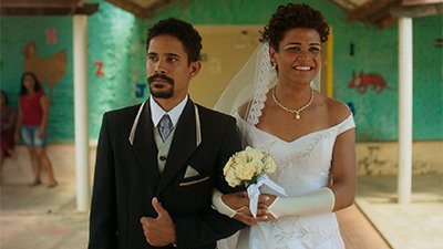 Fotografia do filme Paloma, de Marcelo Gomes, SP, 2022, Ficção, 103 min. Zé, um homem negro, de cabelos curtos, crespos, está de cavanhaque, tem uma fisionomia séria, usa blusa branca, terno preto com detalhes dourados, e gravata cinza. De braços dados com Paloma, mulher trans, negra, cabelos curtos adornados com véu branco, maquiada com sombra dourada e batom vermelho, exibe um largo sorriso. Nas mãos usa luvas e segura um buquê de rosas brancas.