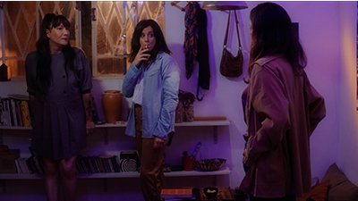 Fotografia de uma cena do filme Tea for two, de Júlia Katharine - SP, 2018, FICÇÃO, 25 min. Três mulheres de pele clara, em uma sala pouco iluminada. De costas para nós, Silvia, cabelos lisos pretos abaixo do pescoço, casaco marrom claro de manga longa com as mãos na cintura, de frente para as outras duas. No meio, com cabelos lisos pretos soltos ao longo do pescoço, camisa branca por baixo da azul clara de manga longa, calça folgada marrom clara, cigarro na boca e com olhar fixo para Silva. Ao seu lado, a terceira, de frente com rosto de perfil direito, pequenos olhos puxados, lábios finos com batom avermelhado, cabelos pretos longos, com franjas, vestido cinza escuro de mangas até os cotovelos. Ao fundo, parede branca, com duas prateleiras de livros, janela de vidro transparente com grades triangulares brancas, e canteadas, no chão, almofadas grandes marrons.