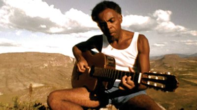 Fotografia de uma cena do filme Tempo rei, de Lula Buarque de Holanda, Andrucha Waddington e Breno Silveira. RJ, 1996, DOCUMENTÁRIO, 107 min., Cor, H264. O jovem cantor Gilberto Gil, homem negro de pele escura, cabelos pretos crespos, em curto black, mais raspados nas laterais, sobrancelhas volumosas pretas, cenho franzido, nariz arredondado de ponta mais fina, orelhas pequenas, lábios grossos em meio bico. Usa camisa regata branca, short azul curto, sentado no topo de um morro, tem a cabeça levemente inclinada para sua direita, olhar fixo para a própria mão esquerda que dedilha no braço do violão. Ao fundo, do cenário, vários morros esverdeados que contrastam com o céu azul claro com densas nuvens brancas.