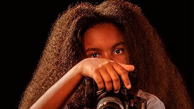 Uma jovem negra retinta, dos ombros para cima, de cabelos crespos, longos, castanhos escuros. Ela tem a testa larga, sobrancelhas grossas, olha para nós por sobre a mão à esquerda, que empunha uma câmera fotográfica profissional. Usa uma camiseta azul clara.