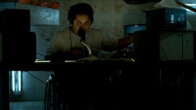 Num cômodo de azulejos florais amarelados, um homem negro, de cabelos curtos, pretos, crespos, olhar contraído, nariz largo, camiseta bege e fone de ouvidos grandes, está à frente de uma mesa, com um microfone. Ele tem o braço à direita com a mão mexendo nos equipamentos de som deste lado. Do outro lado, outras duas caixas, estão escurecidas pela pouca luz, que ilumina mais o rosto dele. Embaixo da mesa, ao fundo, à esquerda, duas luminárias verticais, clareiam a cadeira de rodas que ele senta.