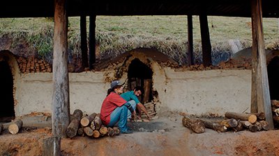 Em frente a um armazém rebaixado, de parede de reboco envelhecido, irregular e esbranquiçado, com toras de madeira empilhadas, uma mulher e um homem, de perfil direito, estão sentados em toras. O homem, próximo a um buraco na parede, com tijolos soltos, é pardo, de cabelos pretos, curtos ondulados e de bigodes pretos. Cabisbaixo, usa uma camisa de manga curta azul e calça azul mais clara, tem o braço direito estendido sobre o joelho. A mulher, um pouco antes dele, é mais clara, está de boné azul, tem os cabelos ondulados, presos num curto rabo de cavalo, usa uma camiseta vermelha, sobre uma de manga comprida azul e está de calça jeans. Também tem o braço direito sobre o joelho e a cabeça está voltada para frente. No entorno deles há outras toras espalhadas, barro e fuligem. Sustentando um teto sombreado, seis vigas de madeira encobrem o espaço que estão. Atrás do armazém segue um descampado gramado.