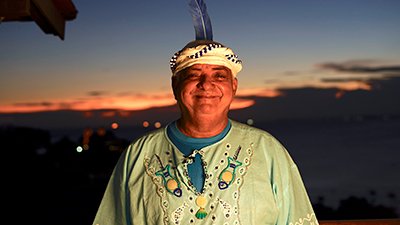 À frente da cidade, durante um por do sol, de céu azul, uma faixa alaranjada e a abaixo, no escuro da noite, luzes de carros e prédios, Gerônimo. Ele usa um turbante branco e azul, enrolado na testa, com uma pena azul presa na parte de trás. Ele é negro não retinto, gordo, o sorriso deixa os olhos encolhidos, usa uma bata verde clara, com bordados no peito, sobre uma camiseta azul.