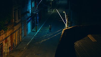 Lá embaixo, no meio de uma rua, à noite, uma pessoa de camisa de manga comprida, amarelada, enrolada até o cotovelo, bermuda azul e calçado, está com os braços ao lado do corpo, estático. Alguns imóveis antigos à esquerda, tem luzes acesas e do outro lado, a luz de um poste reflete no telhado de uma casa, na rua e na pessoa. De um poste a outro emaranhado de fios.