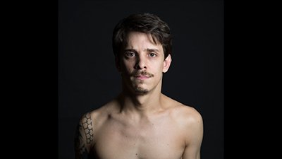 À frente de um fundo preto, um homem branco, do peito para cima. Ele é branco, tem os cabelos castanhos escuros, as sobrancelhas e o bigode castanhos claro, os lábios rosa, cerrados e os olhos focados na gente, caramelo. No ombro à direita, tem uma tatuagem tribal. À esquerda, parte do rosto está sombreado.