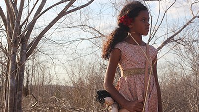 Fotografia de uma cena do filme Rosa Tirana, de Rogério Sagui Num dia de sol e céu azul claro com nuvens brancas, à frente de uma vegetação extremamente seca, um pouco reclinada para a direita, uma menina, de uns 9 anos. Ela é parda, tem os cabelos presos com uma flor vermelha, num rabo de cavalo próximo ao ombro direito. Tem o olhar sério, distante, voltados para a direita e com as sobrancelhas contraídas. Usa um vestidinho de chita, de detalhes rosa e um cinto largo bege. Segura a bolsa a tiracolo rosa, que tem uma boneca de pano pendurada.