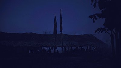 Fotografia de uma cena do filme. Na penumbra noturna, em uma alongada Oca, uma família indigena, em redes e em pé, está reunida. Uma intensa fumaça encobre, acima e o entorno. À direita, algumas palmeiras e aqui na frente duas bandeiras, uma do Brasil.
