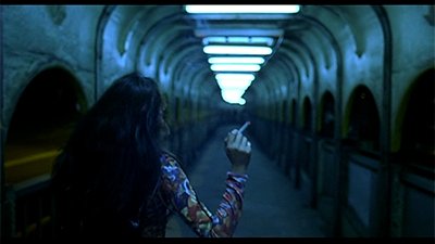 Fotografia de uma cena do filme. Uma mulher, de costas para nós, da cintura para cima, de cabelos lisos, longos, castanhos claro, usa uma blusa colorida, de manga comprida. Tem na mão direita um cigarro. Ela está diante de um longo corredor, com diversas portas arredondadas nas laterais escuras e no alto, lâmpadas de tubo, em sequencia, acesas.