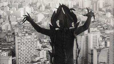 Fotografia em preto e branco de uma cena do filme. Do alto, diante dos edifícios da cidade de São Paulo ocupando todos os espaços, um demônio, de costas para nós. Na contraluz, sombreado, ele tem na cabeça adereço com pontas emendadas, que desce até abaixo da cintura, uma de cada lado. Usa uma roupa justa com uma trança longa na linha da coluna e as mãos abertas além da cabeça, tem os dedos alongados com pontas finas voltados para a cidade.