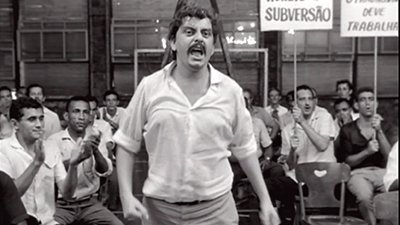 Fotografia em preto e branco, de uma cena do filme. Num galpão, com paredão com janelas pequenas ao fundo, diversos homens sentados, a maioria de camisa social, de manga curta branca e calça, aplaudem e observam um homem que está em pé, bem à frente, no meio. Ele é pardo, tem cabelos curtos encaracolados, tem bigode, usa camisa social, branca de manga comprida arregaçada até os cotovelos e calça social cinza. Ele tem o olhar serio e está com a boca aberta.