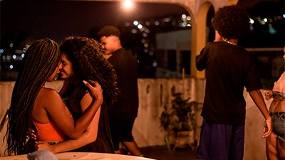 Fotografia de uma cena do filme. Numa varanda, à noite, cinco jovens reunidos. À frente, à esquerda, duas jovens, da cintura para cima dançam com o rosto juntinho. Uma é negra, tem os cabelos longos trançados e está com uma blusa de alça, laranja, ela tem as mãos nos ombros da outra; está é parda, tem um leve sorriso no rosto, os cabelos longos cacheados, está com uma blusa preta e abraça-a pela cintura. Atrás delas, um jovem, quase de costas para nós está próximo à mureta. Ele é negro, tem os cabelos curtos, crespos, pretos e raspados na nuca, olha para esquerda, usa camiseta e bermuda pretas. À direita, outro jovem está de costas, ele tem o cabelo black, preto, usa camiseta preta e bermuda azul marinho. Ao lado dele, uma jovem negro, que só se vê um pedacinho do rosto e parte do corpo. Ela é parda, usa uma blusa rosa e saia jeans. Ao fundo, uma luz amarela impregna o ambiente enquanto pontinhos iluminados atrás distinguem os prédios.