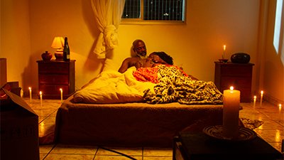 Fotografia de uma cena do filme. Num quarto, deitados numa cama de casal, Zé e uma mulher. Ele é um homem negro, de meia-idade, calvo, de barba grisalha. Está recostado na parede. Em sei peito, uma mulher de cabelos encaracolados, está deitada abraçada a ele. Estão sob cobertores e lençóis branco, vermelho e de oncinha. Acima da cabeceira, uma janela com as cortinas brancas, amarradas. De cada lado da cama, uma mesinha, à esquerda, um pote, um abajur e uma garrafa de vinho, na outra uma vela e um aparelho de som. No chão várias velas acesas e uma de sete dias, sobre um móvel, aqui na frente à direita.