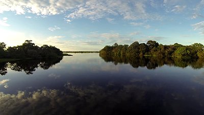 Fotografia de uma cena do filme. Um rio largo aqui na frente e afunilado ao fundo pela vegetação nas laterais, tem refletido no espelho d’água, o céu azul claro, com nuvens esparsas e a suave luz do sol mais intensa à esquerda. O horizonte é uma fina faixa de terra distante.
