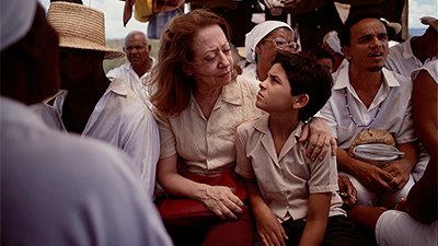Fotografia de uma cena do filme. Sentados, Dora e Josué (Fernanda Montenegro e Vinicius de Oliveira) se olham intensamente, com as sobrancelhas contraídas. Estão entre várias pessoas negras, vestidas de branco, duas mulheres com lenços, um homem com chapéu de palha, outro com uma conta azul e branca. Os dois são brancos. Dora tem os cabelos lisos, ruivos, na altura dos ombros, está com uma camisa de botão, bege, de manga curta, uma calça vinho e segura a bolsa sobre o colo. Josué, Tem os cabelos curtos, lisos, pretos, usa uma camisa de botão, acizentada, de manga curta e um shorts preto. Ele está recostado nela, que o abraça no ombro.