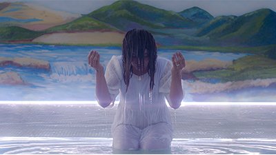 Fotografia de uma cena do filme. Diante de um enorme quadro, com montanhas ao fundo, um lago azul com uma queda d’água que chega ao limite do chão, uma mulher molhada, ajoelhada numa grande poça d’água, vestida de branco, com a cabeça inclinada para a frente. Ela tem os cabelos caídos encobrindo parte da face e escorrendo água e as mãos, ao lado da cabeça voltadas para cima. A água ao seu redor reverbera ondulações e é iluminada pela fresta de luz que saí debaixo do quadro.