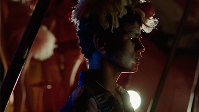 Fotografia de uma cena do filme. Na contra luz, de perfil esquerdo, um adolescente, pardo, de cabelos curtos, crespos, escuro, está com uma peruca curta, loira e uma bola vermelha, acima da testa. As sobrancelhas contraídas e bochecha estão pintadas de branco e o queixo de vermelho. O olhar distante, é marcado por um brilho no olho. Ele usa uma camisa rosa e azul. Atrás, desfocado, o ambiente com duas barras de ferro, uma à esquerda e outra ao fundo à direita, é avermelhado e iluminado por um foco de luz próximo ao ombro esquerdo, que reflete na peruca e clareia o rosto.