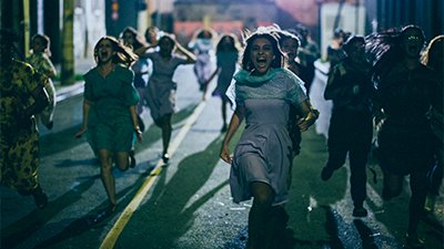 Fotografia de uma cena do filme. Numa rua, à noite, diversas mulheres correm em nossa direção gritando. As do meio usam vestidos azuis e sandálias. Uma delas está com as mãos na cabeça. Muitas delas têm cabelos lisos, abaixo do ombro. Uma luz noturna é irradiada do fundo para frente.
