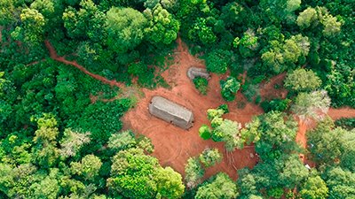 Fotografia de uma cena do filme. Lá embaixo, em meio à floresta, numa manhã de sol, duas ocas. No meio, uma imensa, com cobertura de palha, sobre o chão de terra avermelhada, comprida, tem 3 entradas. Mais acima, um pouco encoberta por árvores pequenas, a outra é bem menor. Mais abaixo, à direita, um cercado e à esquerda, na direção da entrada, uma longa trilha, com uma bifurcação no alto à esquerda.